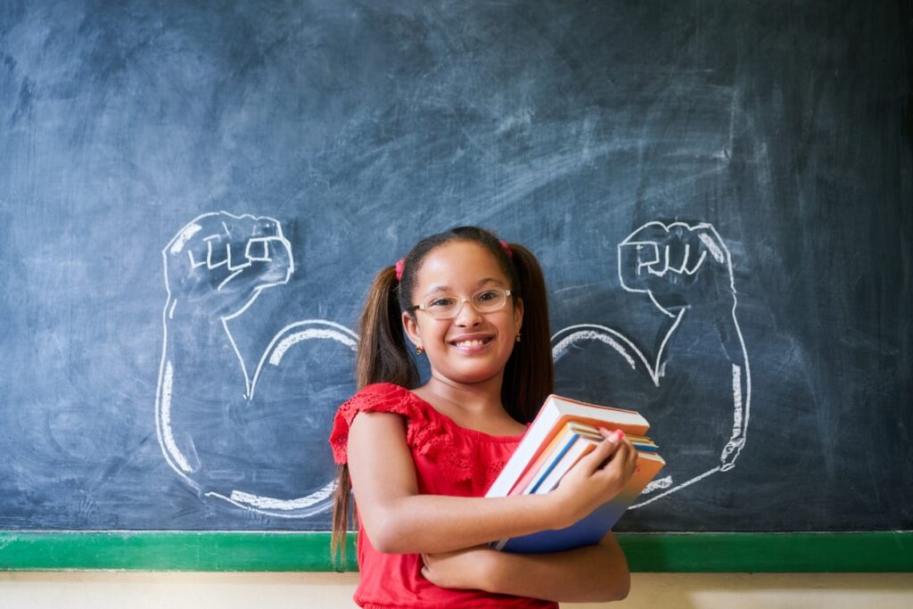 A girl wearing glasses and holding onto a pile of books, smiling for the camera in front of a blackboard