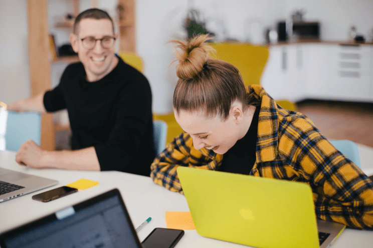 A man and a woman laughing while sitting on a desk with laptops in front of them