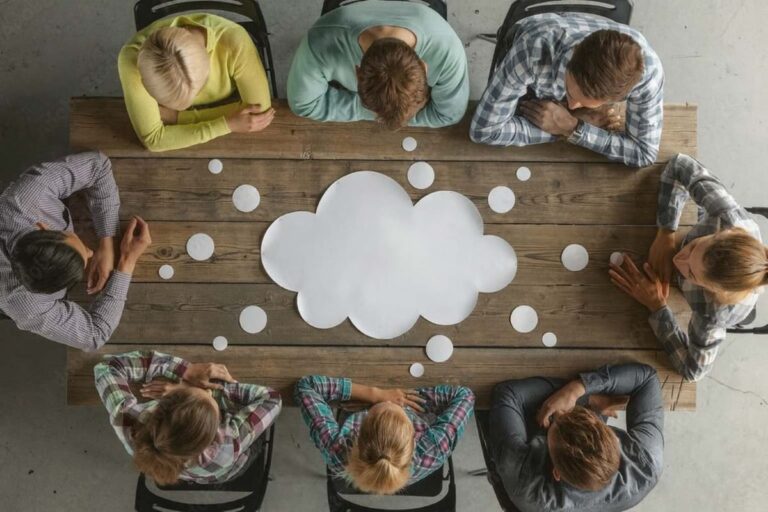 Overhead view of a diverse team in a brainstorming session around a wooden table with a thought cloud in the center, symbolizing leadership dialogue and collaborative problem-solving.